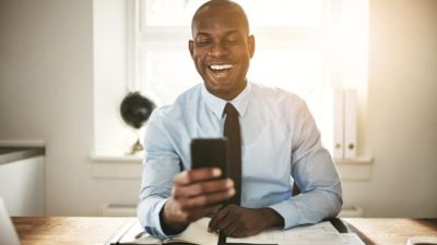 a man sits at his desk wearing a business shirt and tie and has a hearty laugh at something on his mobile phone.