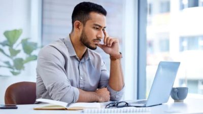 A man in his 30s with a clipped beard sits at his laptop on a desk with one finger to the side of his face and his chin resting on his thumb as he looks concerned while staring at his computer screen.