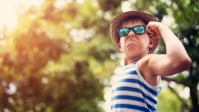 A young boy wearing a hat, sunnies and striped singlet looks fierce and flexes his arm in victory.