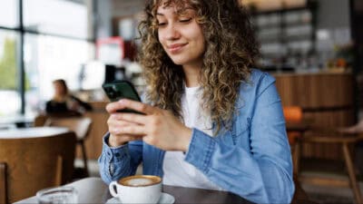 A young woman drinking coffee in a cafe smiles as she checks her phone.