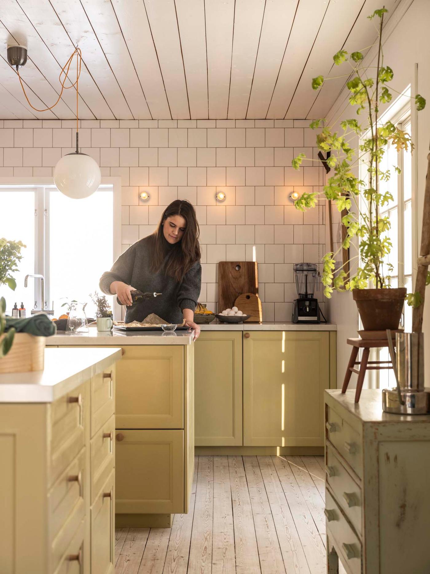 A person stands at an island in the centre of the kitchen with yellow cabinets, holding a bottle of olive oil.