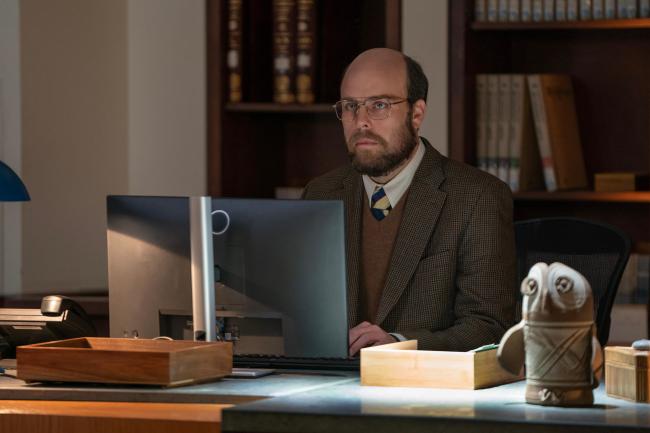 A man sitting at a desk and working on a large laptop, in front of a large bookshelf; still from 'Paradise'