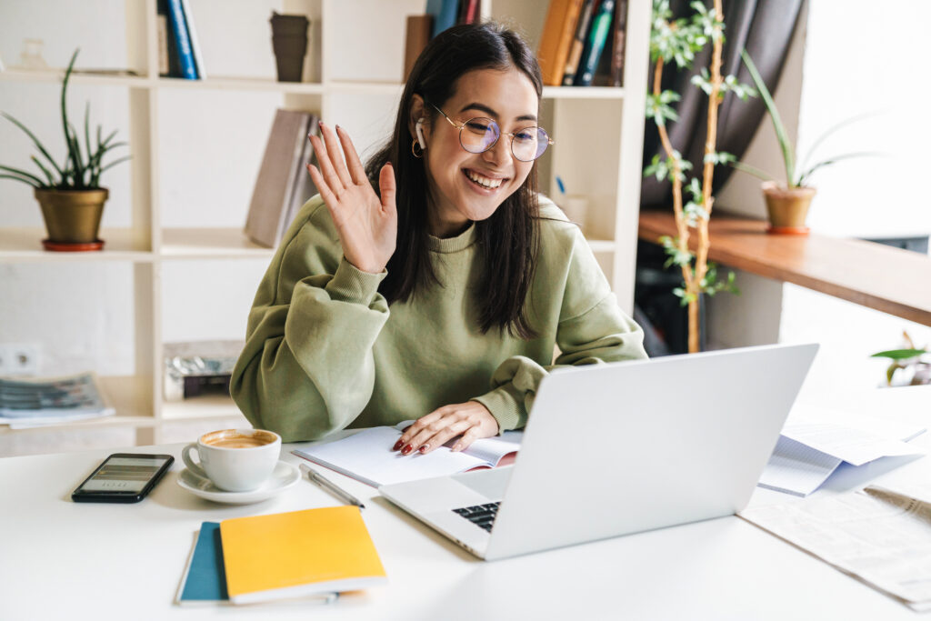 A person working remotely and waving to a laptop screen while in a virtual meeting. 