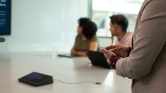 Two women and one man join an in-person meeting in a medium sized conference room with a companion device. One woman is casting from her mobile device while standing at the back of the room.