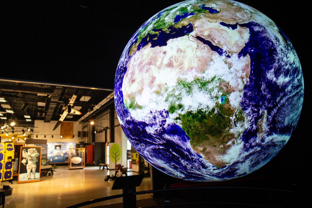 A large sphere with moving satellite images of the Earth projected on it. The sphere shows a brown and green image of the continent of Africa surrounded with blue water and partly obscured by swirling white clouds.