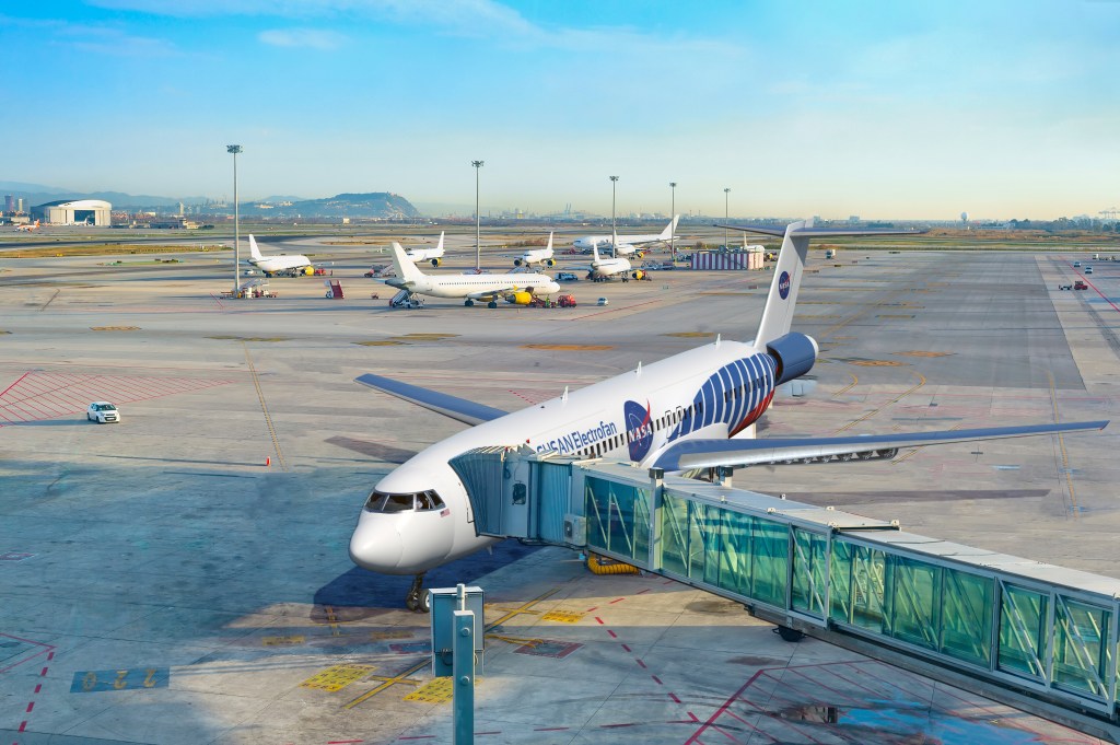 A rendering showing a single-aisle aircraft with a hybrid propulsion system, including distributed wing-mounted electric engines and a single aft engine, parked at an airport gate with a jet bridge extended to the front of the plane. The words “SUSAN Electrofan” and “PAX 180” are displayed along the side of the fuselage, along with the NASA insignia on the side and tail of the aircraft. Several other aircraft sit scattered in the background on the tarmac, with various buildings and hills in the distance.