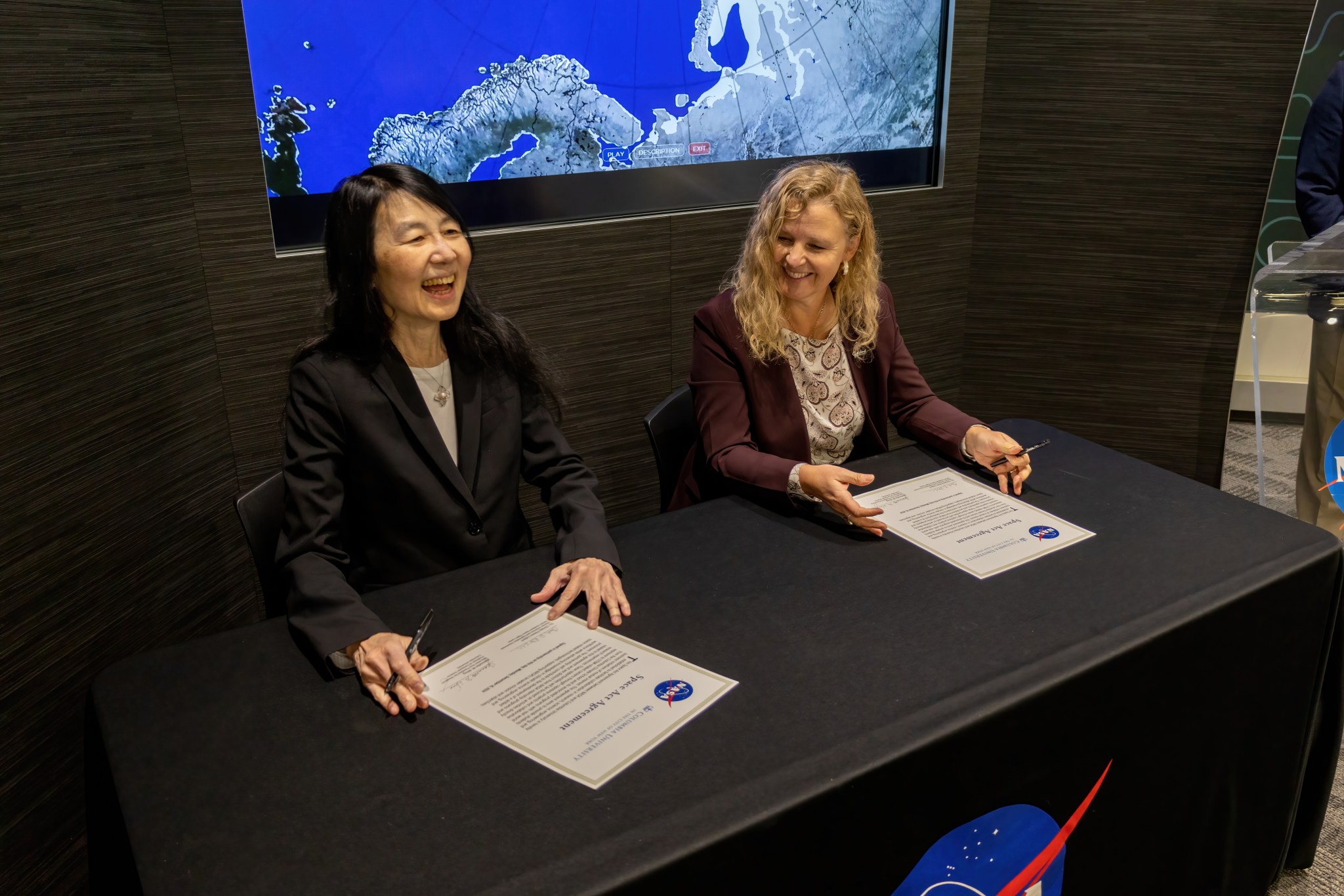 Jeanette Wing and Christa Peters-Lidard sit at a table draped with a black tablecloth with the NASA logo. They are both holding pens and large white documents with the NASA meatball logo sit in front of each of them. They are mid-conversation and both smiling.