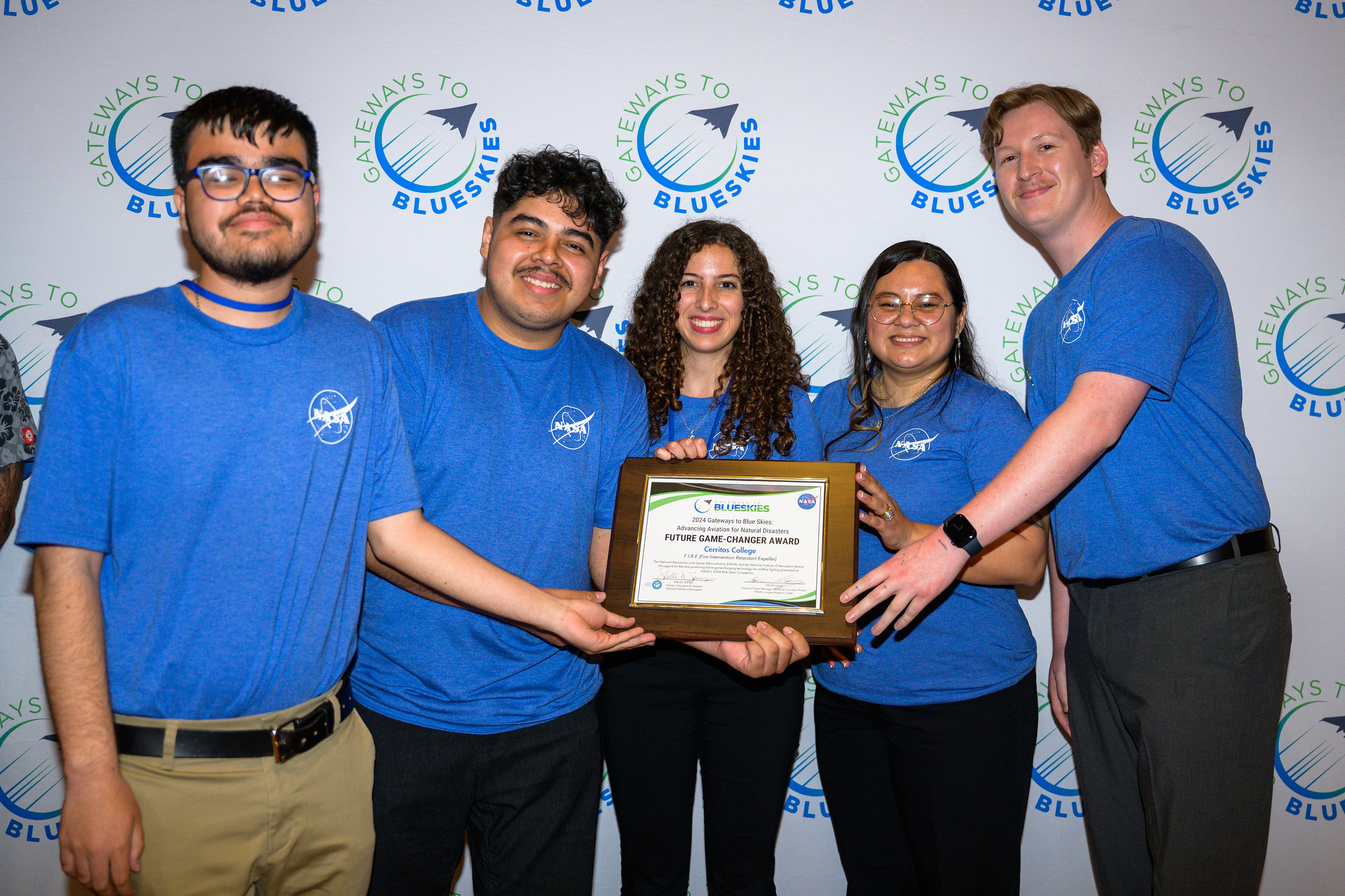 Four people in blue shirts holding a certificate pose in front of a white wall with logos.