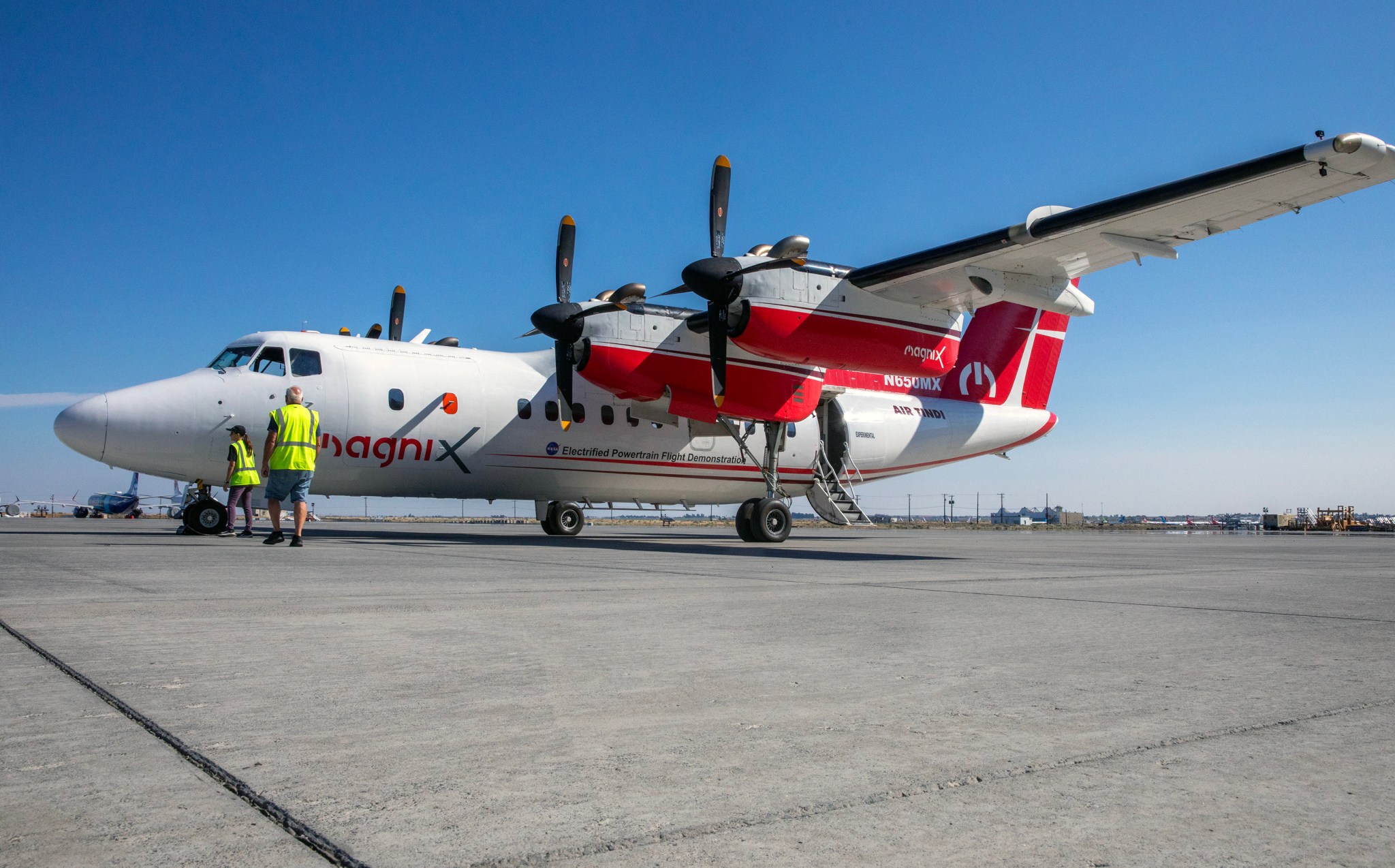 A turboprop plane with a red and white livery sits on a tarmac with a bright blue sky in the background. A man and a woman wearing safety vests stand at the front of the plane.