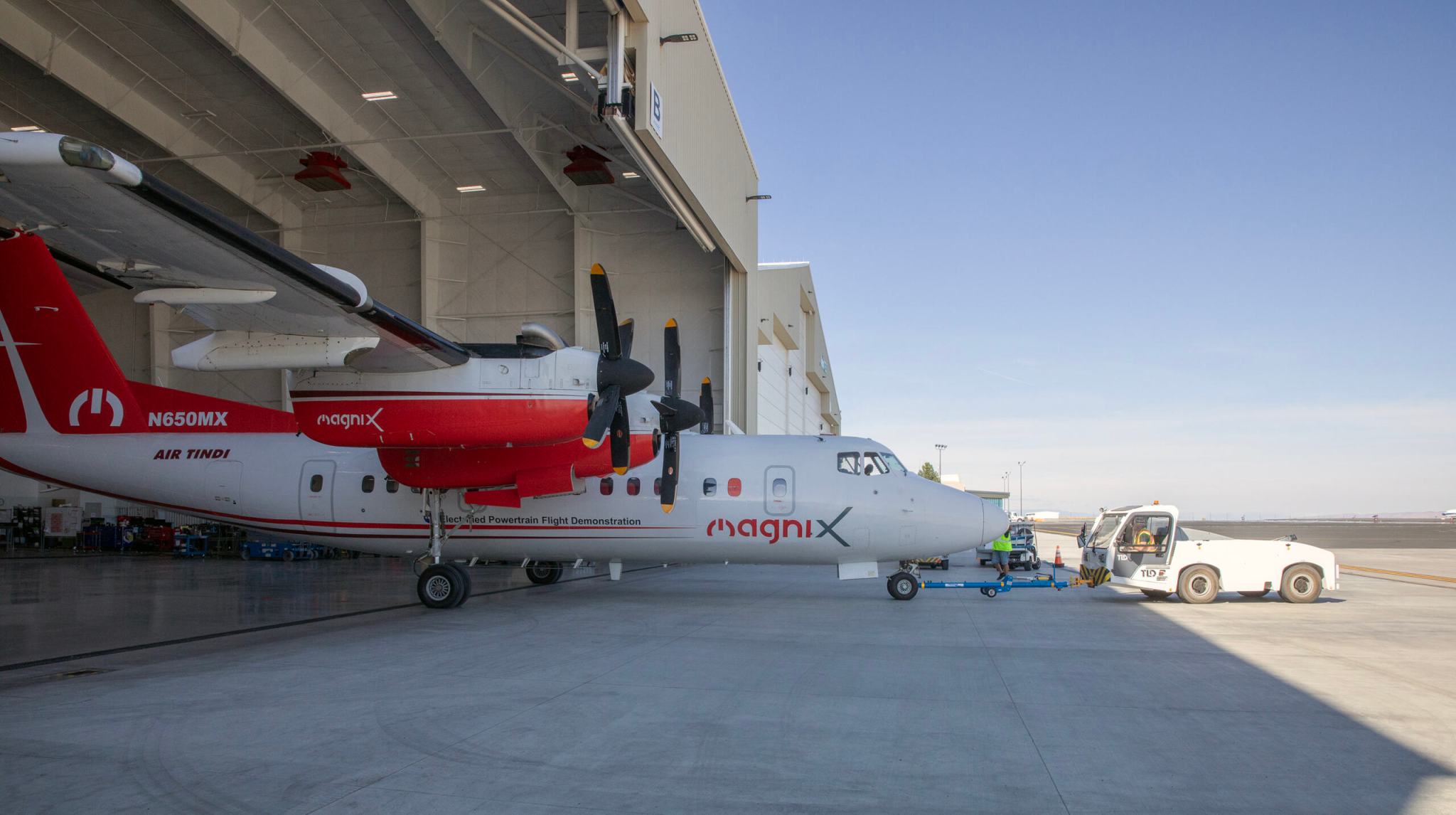 A turboprop airplane with a white and red livery gets towed out of an aircraft hangar with a white truck onto a concrete tarmac with a clear blue sky in the background.