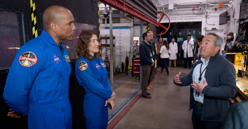 Two astronauts in blue jumpsuits stand at the left of the image, listening to a man speaking on the right side of the image. People and equipment fill the background.
