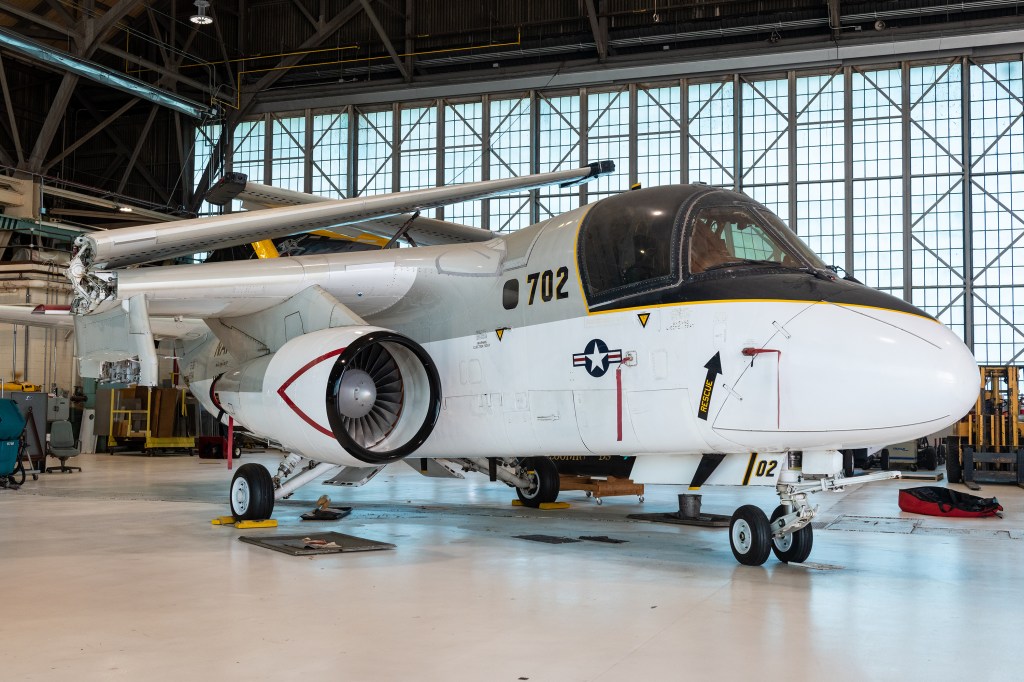 The S-3B Viking aircraft rests in the NASA Glenn Hangar. The plane is painted gray, black, and white. The wings are folded atop the aircraft.