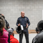 A police officer stands and speaks to a seated group of people in a room with cinder block walls, holding a helmet and papers.