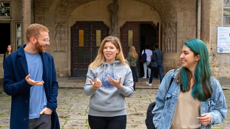 Three students are chatting in front of the main university building.