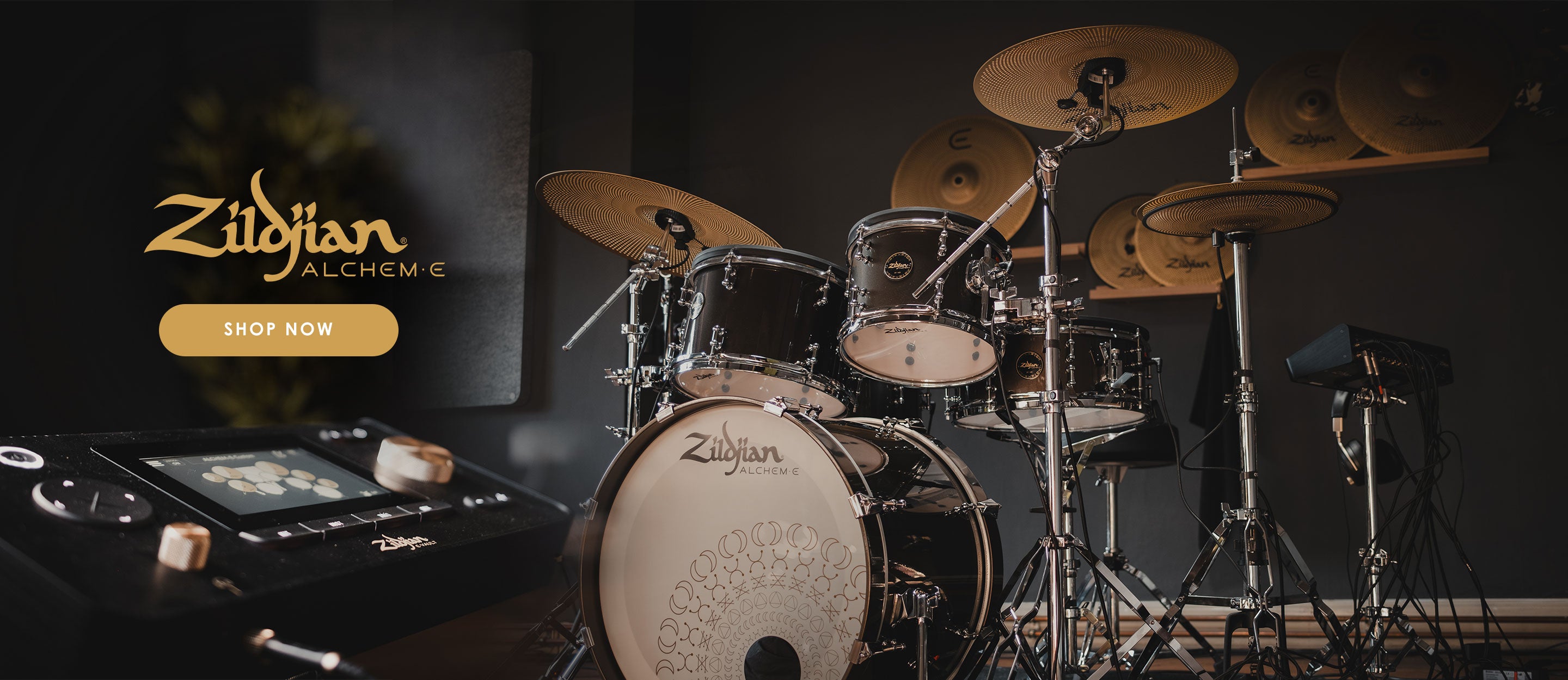 Close-up of a Zildjian Alchem-E electronic drum kit with gold cymbals, black drum shells, and a decorated bass drum head. A touch-screen drum module and cables are visible, with a “Shop Now” button and Zildjian Alchem-E logo prominently displayed.